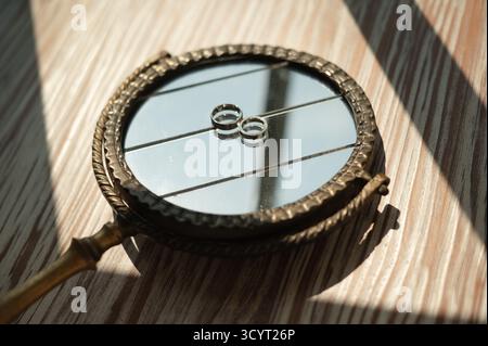 Uno specchio a mano vintage che riflette due fedi nuziali, collocato su una superficie in legno con luce naturale soffusa Foto Stock