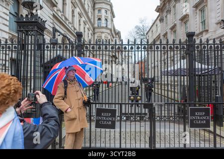 Un uomo con un ombrello della Union Jack si posa fuori dal cancello di Downing Street a Londra, mentre un altro lo fotografa vicino a cartelli, dissuasori e personale di sicurezza. Foto Stock