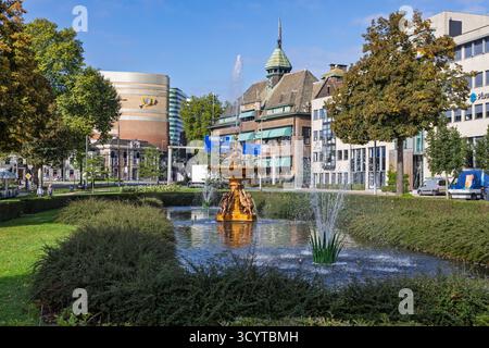 La fontana dorata di Janspark, Arnhem, circondata dal verde e da uno stagno, con edifici storici e moderni sullo sfondo. Foto Stock