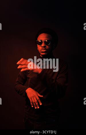 Ritratto di un cantante hip-hop afroamericano con occhiali da sole neri che rappano e gesticolano in studio scuro con luce debole Foto Stock
