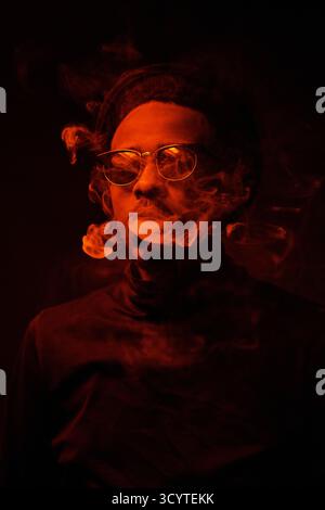Ritratto in vita dell'uomo nero sicuro con eleganti occhiali da sole che esaltano il fumo di sigaretta nella camera buia Foto Stock