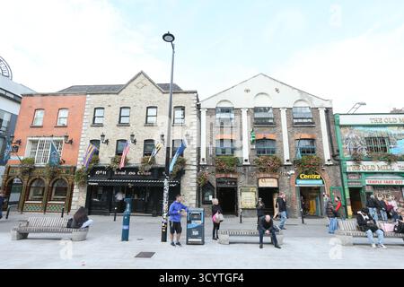 Dublino, Irlanda - 1 ottobre 2025 - Una fila di pub e ristoranti visti da Temple Bar Square mentre la gente siede sulle panchine della città di Dublino in un pomeriggio di sole raffigurante una scena di strada nella capitale irlandese Foto Stock