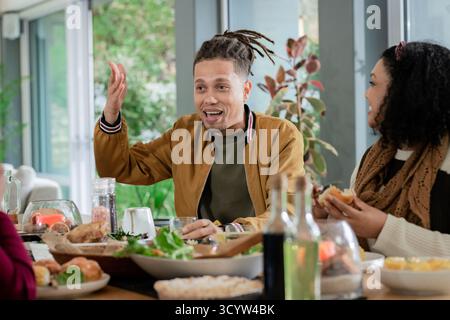 Diversi amici condividono il pranzo nella sala da pranzo di casa intorno a un tavolo di legno con insalata e pasticcini Foto Stock