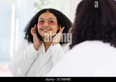 Donna adulta che regola le patch in idrogel sotto gli occhi sullo specchio di cortesia indossando un accappatoio bianco Foto Stock