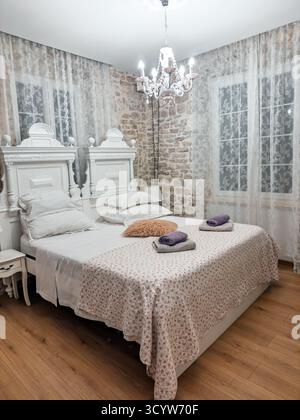 Camera incantevole e accogliente con un'estetica vintage, dotata di un classico letto bianco con copriletto floreale, parete in pietra a vista, elegante lampadario e. Foto Stock