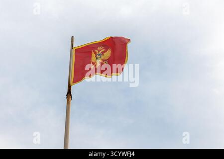 Bandiera del Montenegro con un bordo dorato onde da un palo di legno contro un cielo blu brillante. L'emblema presenta un'aquila e una corona che simboleggiano il cerimoniale Foto Stock