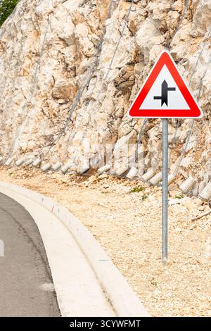 Un segnale di avvertimento triangolare si trova accanto a una strada curva su un pendio roccioso, protetto da reti. La scena trasmette consapevolezza dei pericoli, sicurezza stradale e. Foto Stock