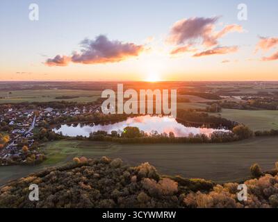 il lago still rispecchia il cielo scintillante del tramonto, circondato da alberi autunnali, campi aperti e un tranquillo villaggio immerso nel tranquillo paesaggio rurale Foto Stock