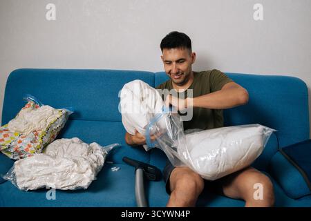 Un giovane sorridente che confeziona cuscini bianchi e biancheria da letto in sacchetti sottovuoto seduti sul divano blu del soggiorno, pronti a muoversi o a riordinare Foto Stock