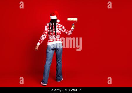 Una divertente pittrice delle feste con un maglione natalizio dipinge una parete rossa con un rullo mentre una donna sorridente indossa il cappello e i jeans di Babbo Natale Foto Stock