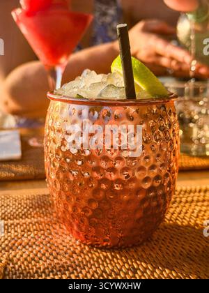 Cocktail mulo moscovita in una tazza di rame martellata con fetta di lime e paglia su un tappeto intrecciato, con persone in sottofondo che si godono delle bevande Foto Stock
