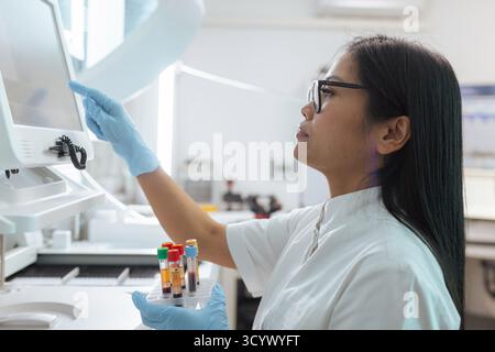 Ricercatrice di laboratorio che tiene in mano le provette per analisi del sangue e regola un analizzatore biochimico automatico. Foto Stock