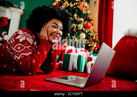 La donna sorridente in maglione rosso ama le videochiamate durante le festività natalizie vicino a alberi decorati e regali luminosi. Foto Stock