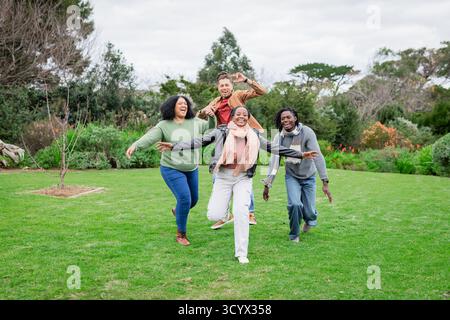 Diversi amici che corrono giocosamente sul prato verde del parco indossando giacche e sciarpe Foto Stock