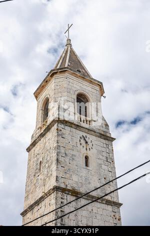 La storica torre della chiesa in pietra si erge con una croce, adagiata su un cielo blu parzialmente nuvoloso. Architettura rustica, torre a cupola e vegetazione circostante Foto Stock