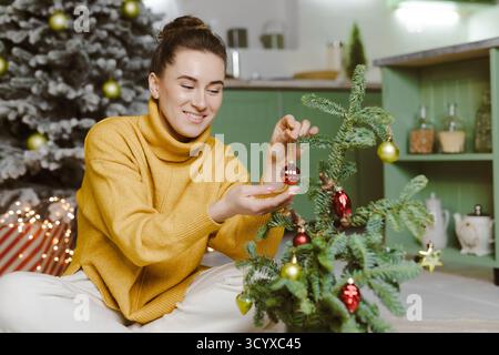 Donna sorridente in un caldo maglione decora un piccolo albero in una cucina accogliente, ideale per annunci di vacanza, saluti, negozi e storie di stile di vita. Foto Stock