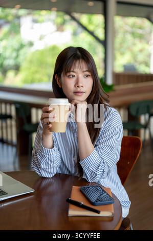 Donna asiatica in camicia blu con una mano sotto il mento che tiene una tazza di caffè mentre siede con un computer portatile al tavolo di legno nel caffè. Studiare o lavorare all'esterno. Foto Stock
