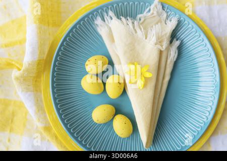 Questo vivace tavolo pasquale presenta un piatto blu adornato con uova gialle e un delicato fiore. La tovaglia a scacchi aggiunge un tocco natalizio Foto Stock