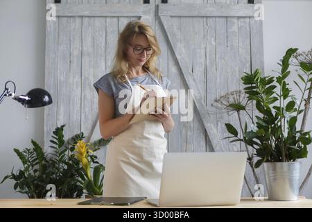 La fiorista ragazza in occhiali e grembiule scrive su un taccuino. Concetto floreale Foto Stock