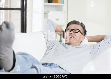 Uomo asiatico maturo anni '50 che sogna ad occhi aperti e riposa a casa. Un uomo cinese anziano rilassato e disteso su un divano interno Foto Stock