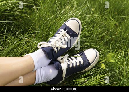 L'immagine raffigura i piedi di una persona che indossa scarpe da ginnastica blu e calzini bianchi, appoggiati su una vibrante erba verde sotto la luce del sole Foto Stock