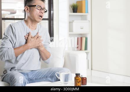 Uomo asiatico maturo degli anni '50, che preme sul petto con espressione dolorosa, seduto sul divano a casa, medicine sul tavolo Foto Stock