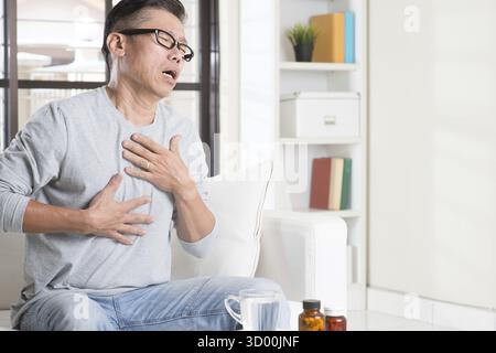 Ritratto di un uomo asiatico maturo degli anni '50, che preme sul petto con espressione dolorosa, seduto sul divano a casa, medicine e acqua sul tavolo Foto Stock