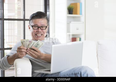 Ritratto di uomo asiatico maturo felice anni '50 che usa il computer Internet e conta i soldi a casa, guadagnando soldi dal suo business online di successo. Lavoro f Foto Stock