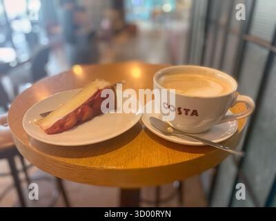 Burgas, Bulgaria - 26 settembre 2025: La tazza di caffè Costa piena di cremosa latte art è collocata accanto a una fetta di torta su un tavolo di legno nell'accogliente caffè, cr Foto Stock