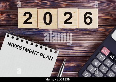 Concetto di pianificazione finanziaria. Blocco note con testo del piano di investimento, blocchi in legno con 2026 e Calcolatrice su sfondo di una scrivania in legno. Rendering 3d. Foto Stock