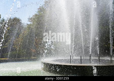 Elegante fontana circolare con alto getto centrale e ruscelli d'acqua circostanti in un parco verde sotto il cielo azzurro, a simboleggiare il relax e la vita urbana Foto Stock