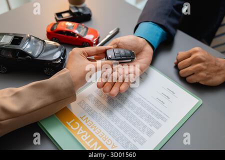 Un uomo d'affari e un cliente stanno esaminando e firmando un contratto di vendita di auto presso una scrivania, con modellini di auto di piccole dimensioni, una tazza di caffè e documenti intorno a loro, Foto Stock