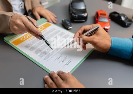 Un uomo d'affari e un cliente stanno esaminando e firmando un contratto di vendita di auto presso una scrivania, con modellini di auto di piccole dimensioni, una tazza di caffè e documenti intorno a loro, Foto Stock