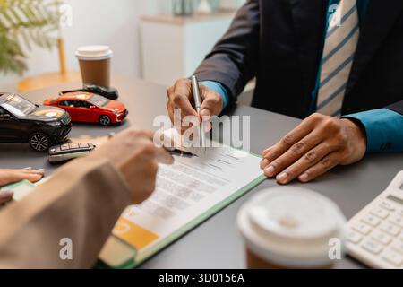 Un uomo d'affari e un cliente stanno esaminando e firmando un contratto di vendita di auto presso una scrivania, con modellini di auto di piccole dimensioni, una tazza di caffè e documenti intorno a loro, Foto Stock