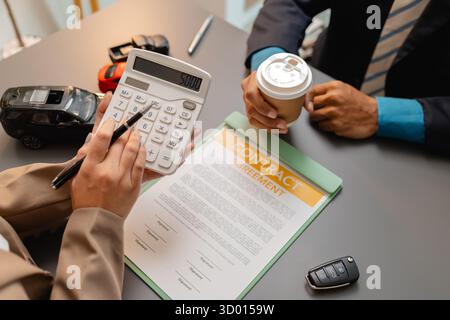 Un uomo d'affari e un cliente stanno esaminando e firmando un contratto di vendita di auto presso una scrivania, con modellini di auto di piccole dimensioni, una tazza di caffè e documenti intorno a loro, Foto Stock