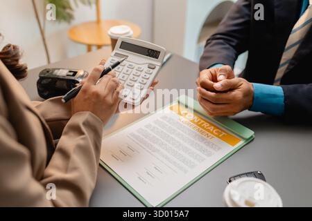 Un uomo d'affari e un cliente stanno esaminando e firmando un contratto di vendita di auto presso una scrivania, con modellini di auto di piccole dimensioni, una tazza di caffè e documenti intorno a loro, Foto Stock