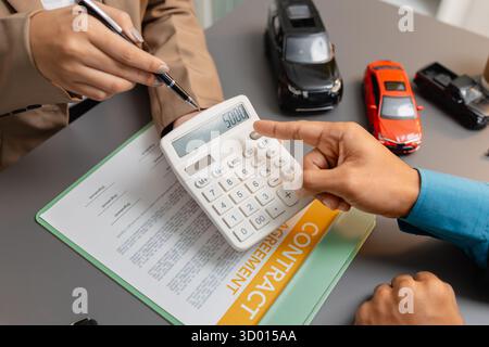 Un uomo d'affari e un cliente stanno esaminando e firmando un contratto di vendita di auto presso una scrivania, con modellini di auto di piccole dimensioni, una tazza di caffè e documenti intorno a loro, Foto Stock