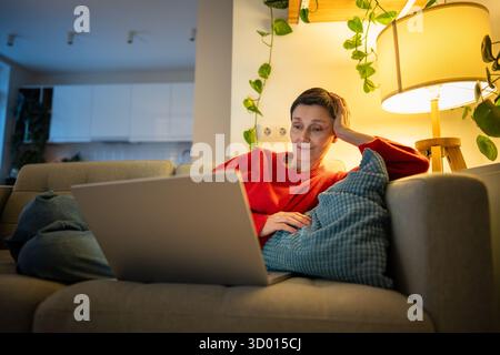 Una donna sorridente e felice sdraiata sul divano guardando film sul laptop in un salotto accogliente da sola, riposando le donne Foto Stock