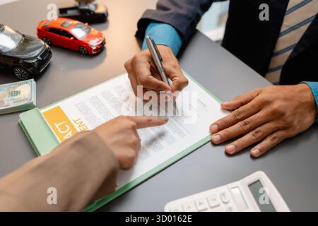 Un uomo d'affari e un cliente stanno esaminando e firmando un contratto di vendita di auto presso una scrivania, con modellini di auto di piccole dimensioni, una tazza di caffè e documenti intorno a loro, Foto Stock
