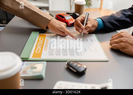 Un uomo d'affari e un cliente stanno esaminando e firmando un contratto di vendita di auto presso una scrivania, con modellini di auto di piccole dimensioni, una tazza di caffè e documenti intorno a loro, Foto Stock