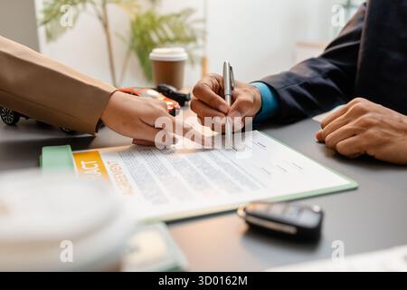Un uomo d'affari e un cliente stanno esaminando e firmando un contratto di vendita di auto presso una scrivania, con modellini di auto di piccole dimensioni, una tazza di caffè e documenti intorno a loro, Foto Stock