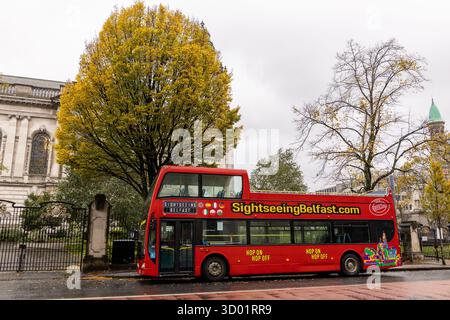 Autobus turistico rosso a due piani parcheggiato in una strada piovosa a Belfast in autunno con alberi ed edifici storici sullo sfondo Foto Stock