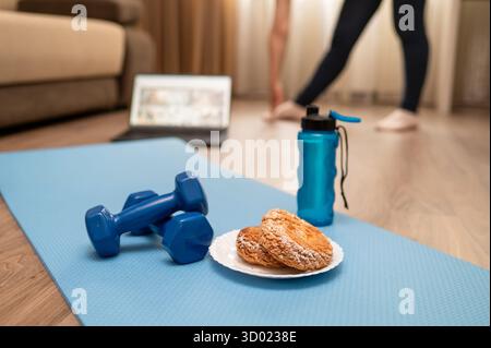 Attrezzature fitness con manubri, rullo in schiuma, bottiglia d'acqua, cuffie e piatto di biscotti sul tappetino yoga a casa, laptop e gambe femminili sullo sfondo Foto Stock