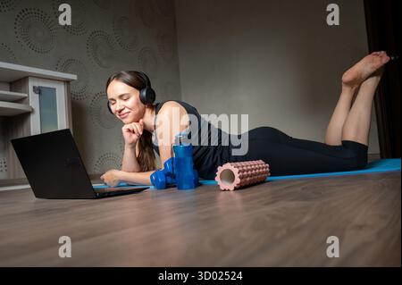 Giovane donna che guarda l'allenamento online sul laptop mentre si sdraia su un tappetino da yoga con le cuffie, accanto a manubri, bottiglia d'acqua e rullo in schiuma, concetto di hom Foto Stock