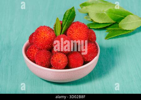 Frutti di arbutus in una piccola ciotola rosa, con due foglie verdi in piedi, il frutto di Arbutus unedo noto anche come albero di fragola, rosso vivo e. Foto Stock
