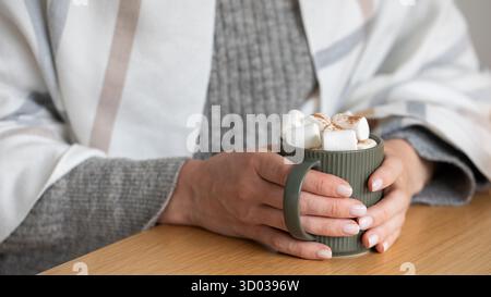 Donna matura in maglione lavorato a maglia con plaid caldo tiene la tazza di cacao al primo piano della scrivania di legno. Lady scalda le mani con il drink nel periodo freddo dell'anno. Stile di vita accogliente Foto Stock