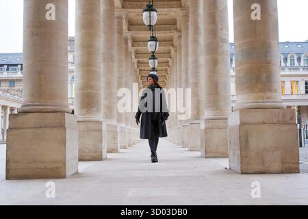 Una donna alla moda in abbigliamento invernale cammina tra le colonne di pietra del Palais Royal. Il design simmetrico e lo stile parigino creano una scena senza tempo ed elegante. Foto di alta qualità || modello rilasciato Foto Stock