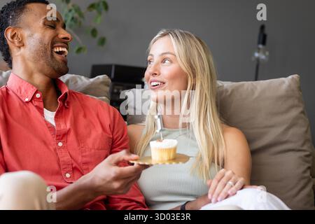 Coppia che ride e si diverte con il cupcake insieme sul comodo divano di casa. Relax, dessert, legame, stile di vita, interni, felicità || modello rilasciato Foto Stock