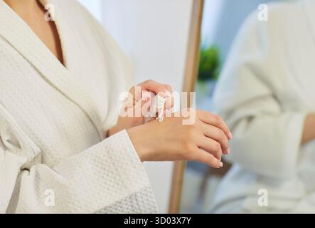 Donna bionda in accappatoio che applica crema per le mani dopo la doccia, specchio spa riflesso Foto Stock
