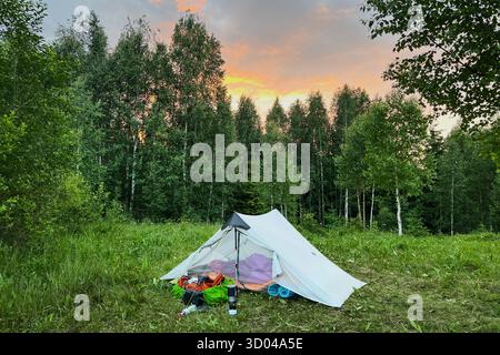 Tenda bianca per turisti con attrezzatura da campeggio in radura erbosa sullo sfondo di fitti alberi. Il cielo risplende di vivaci sfumature di arancione e rosa al tramonto, creando un'atmosfera serena sul tranquillo campeggio. Foto Stock